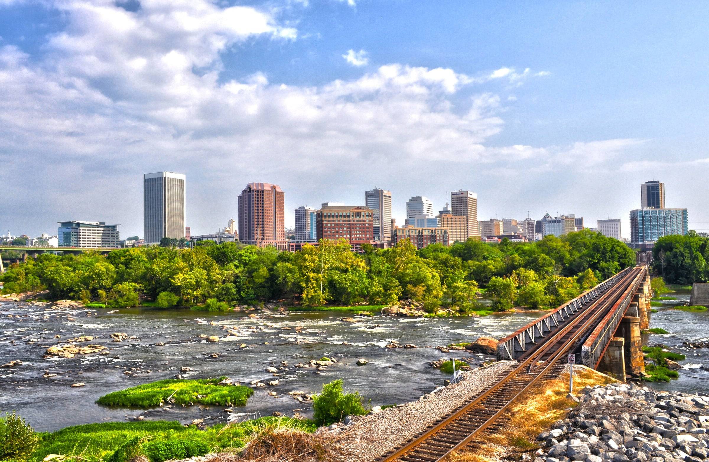 Downtown Richmond skyline above the rocky rapids of the James River, with a railroad trestle along the right bank